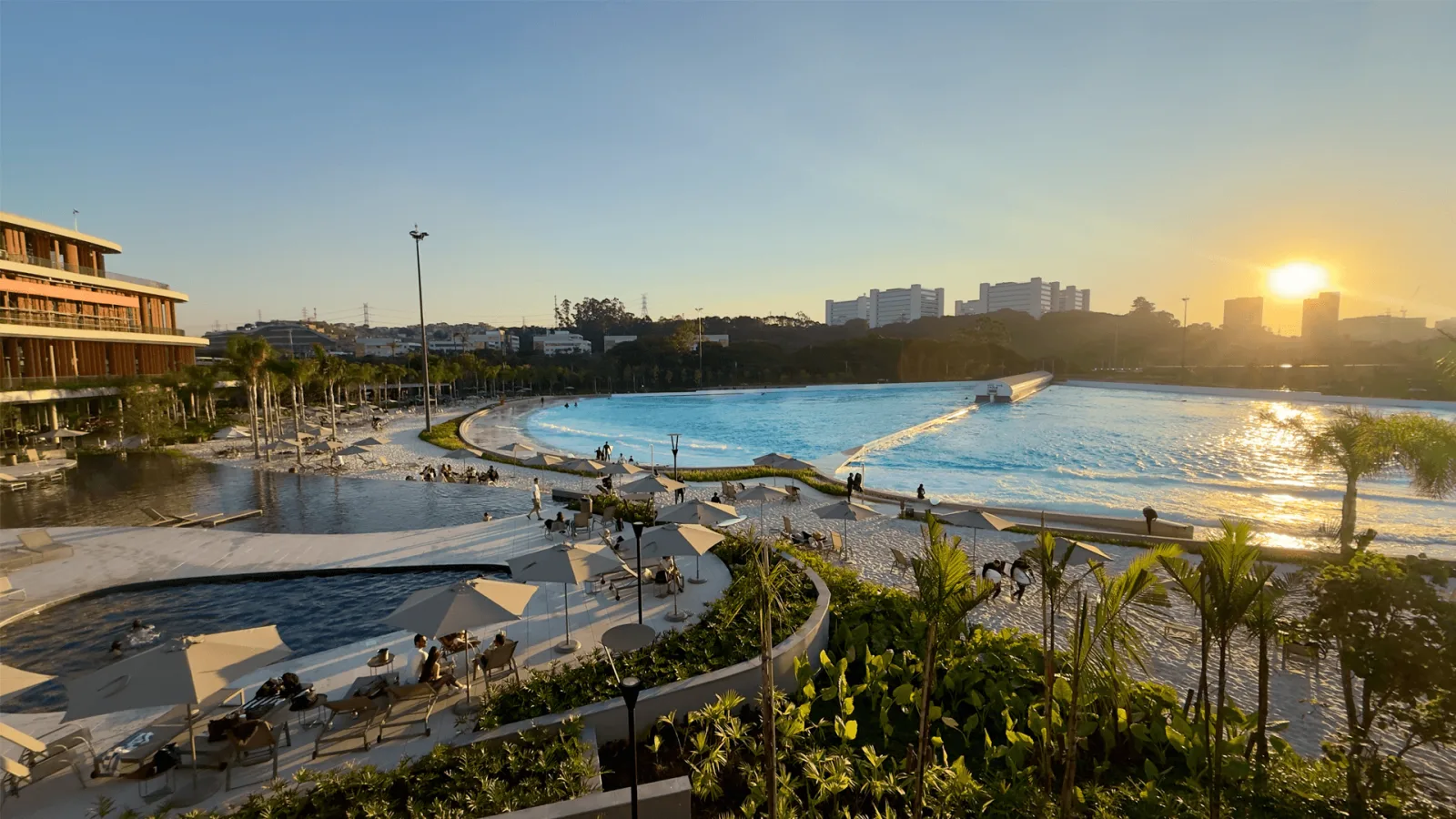 Pôr do sol no Beyond The Club, com a piscina de ondas, a praia de areia branca, o prédio do clube à esquerda e os guarda-sóis entre os jardins