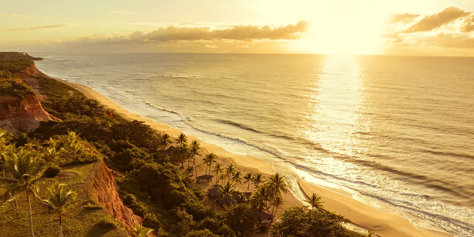 Falésias avermelhadas e coqueiros à beira da praia ao entardecer, com o sol refletido no mar