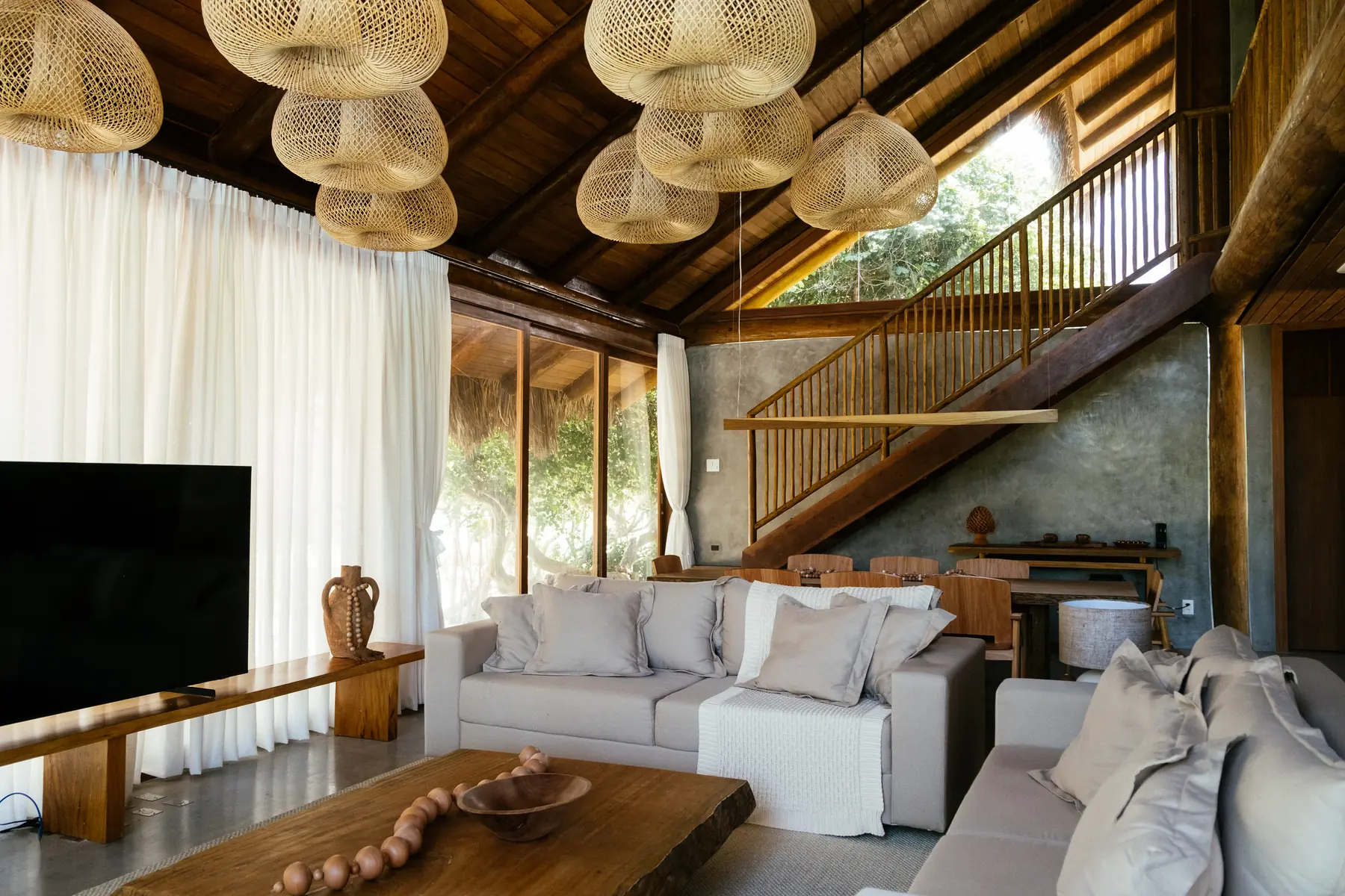 Sala de estar de uma casa em madeira e palha, com pé-direito alto, escada e luminárias de fibra